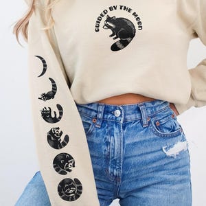 May include: A sand-colored crewneck sweatshirt featuring a black graphic of a raccoon and the text "GUIDED BY THE MOON" on the chest. The right sleeve displays a vertical sequence of five black raccoon illustrations in various poses.