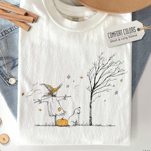 Può includere: Una t-shirt bianca con un'illustrazione a linee nere di uno spaventapasseri con cappello giallo, una zucca, un cane e un albero spoglio con foglie che cadono. Un'etichetta sulla maglietta riporta COMFORT COLORS Short & Long Sleeve.