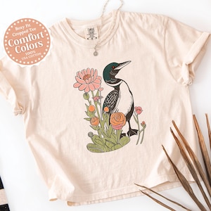 May include: Ivory-coloured, boxy fit cropped tee with a graphic of a bird and flowers. The bird is black, white, and green, and the flowers are pink and orange. The shirt has rolled-up sleeves and the words "Comfort Colors" are printed on a circular label.