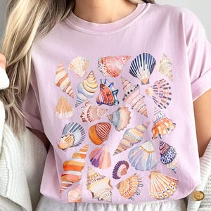 Könnte beinhalten: Hellrosa T-Shirt mit einem Aquarelldesign verschiedener Muscheln in Orange, Blau, Rosa und Weiß. Das Shirt hat kurze Ärmel und einen Rundhalsausschnitt. Das Wort "Blossom" ist unten links sichtbar.