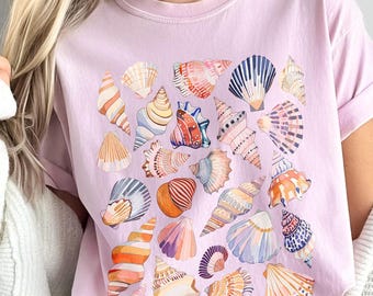 Op de afbeelding: Een lichtroze t-shirt met een aquarelontwerp van diverse schelpen in oranje, blauw, roze en wit. Het shirt heeft korte mouwen en een ronde hals. Het woord "Blossom" is linksonder zichtbaar.