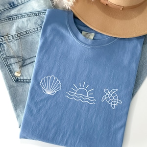May include: A light blue t-shirt with white seashell, sun, and turtle graphics, paired with light wash denim jeans and a tan hat. A gold necklace is visible. The shirt is labeled "Washed Denim".