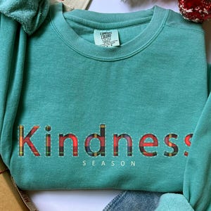 May include: A teal sweatshirt with the word "Kindness" in a plaid pattern across the chest. The word "Season" is below the word "Kindness". The sweatshirt is folded with the sleeves showing. A Christmas-themed background is visible.