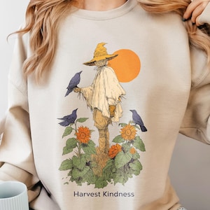 May include: A sand-colored crewneck sweatshirt featuring a graphic illustration of a scarecrow wearing a yellow hat, surrounded by sunflowers and three black crows, with a large orange sun and the text Harvest Kindness printed below.