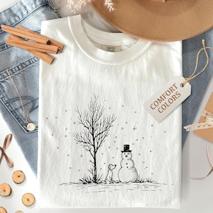 以下が含まれることがあります： 雪だるま、犬、木が雪景色の中に描かれた白のComfort Colors Tシャツ。Tシャツには「Comfort Colors」と書かれたタグが付いています。