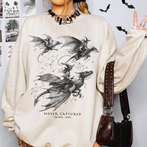 May include: A sand-colored crewneck sweatshirt featuring a black ink-style graphic of figures riding dragons. The text reads NEVER CAPTURED SALEM-1692. The design includes detailed line work of wings and stars, printed on a long-sleeved pullover garment.