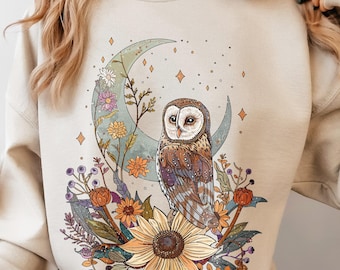 Pode incluir: Uma camisola de gola redonda cor de areia com uma impressão gráfica de uma coruja castanha pousada numa lua crescente rodeada de flores coloridas, incluindo um girassol grande, com a palavra SAND impressa no canto superior esquerdo.