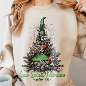 May include: A sand-colored crewneck sweatshirt featuring a graphic of a green witch hat styled as a Christmas tree, decorated with red ornaments, white poinsettias, and a crescent moon. The text reads Our Spirit Remains - Salem 1692 -.
