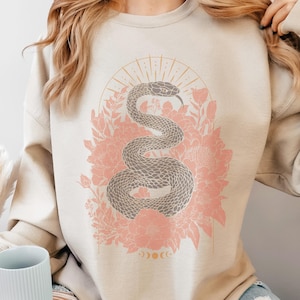 Può includere: Una felpa beige con una stampa grafica di un serpente avvolto attorno a fiori rosa. Il serpente è grigio scuro e i fiori sono rosa chiaro. La grafica è circondata da un'esplosione di sole dorata e una luna crescente.