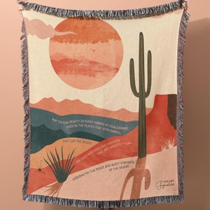 May include: A woven tapestry throw blanket with a desert landscape design. The blanket features a large red sun, a tall green cactus, and a quote about finding beauty in every corner of your journey, just like the desert. The quote reads: "May you find beauty in every corner of your journey, even in the places that seem barren, just like the desert. You hold mysteries and wonders that are endlessly yours. Wishing you the peace and quiet strength of the desert."