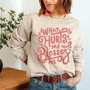 May include: A beige sweatshirt with a pink graphic that says "What Hurts Me Blesses Me". The person wearing the sweatshirt is also wearing blue jeans and a brown belt.