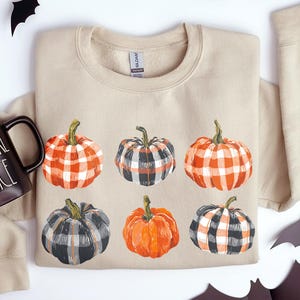 May include: Sand-colored sweatshirt featuring six watercolor pumpkins in various plaid patterns. A black mug with "VAMPIRE JUICE" text sits to the left. Black bat cutouts are scattered around the sweatshirt, creating a Halloween theme.