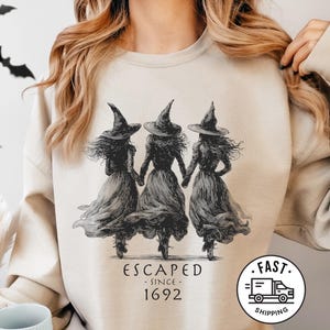 Könnte beinhalten: Beiges Sweatshirt mit einer Schwarz-Weiß-Illustration von drei Hexen, die sich an den Händen halten. Der Text "ESCAPED SINCE 1692" ist unter dem Bild aufgedruckt. Das Sweatshirt hat einen Rundhalsausschnitt und lange Ärmel.