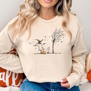 Pode incluir: Uma camisola de gola redonda cor de areia com uma ilustração de linhas pretas de um espantalho, um cão pequeno, uma abóbora e uma árvore sem folhas. O design inclui detalhes subtis em laranja na abóbora e nas folhas no centro do peito.