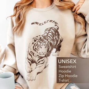 Op de afbeelding: Een zandkleurige sweater met ronde hals en een grote, donkerbruine grafische illustratie van een tijger in een springende houding. De tekst op de afbeelding luidt: Sand, UNISEX, Sweatshirt, Hoodie, Zip Hoodie, T-shirt.