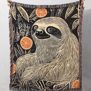 Puede incluir: Una manta tejida en blanco y negro con un diseño gráfico de un perezoso y naranjas. El perezoso está en una pose relajada con los brazos cruzados. Las naranjas están dispersas alrededor del perezoso.