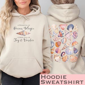 Peut inclure: Un sweat à capuche beige avec l'inscription "A pocketful of ocean magic" et "A heart full of joy and wonder" sur le devant. Le dos du sweat est orné d'un motif de coquillages colorés.
