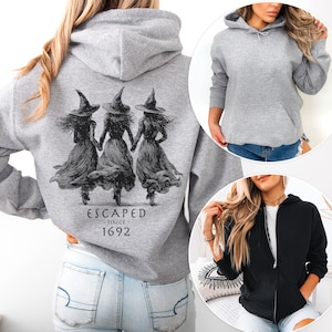 Puede incluir: Sudadera con capucha gris deportivo con un gráfico de tres brujas tomadas de la mano, con el texto "ESCAPED SINCE 1692" impreso en la espalda. Disponible como sudadera, sudadera con capucha y sudadera con cremallera.
