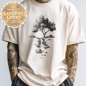 May include: Ivory-coloured t-shirt with a black and white tree graphic. The shirt has a round, gold-coloured emblem that reads "Unisex Vintage Style Comfort Colors 100% Cotton". The word "Ivory" is printed at the bottom of the shirt.