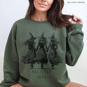 May include: A military green crewneck sweatshirt featuring a black graphic of three witches in pointed hats holding hands. Below the illustration, the text reads ESCAPED - SINCE - 1692. The garment has long sleeves and a ribbed neckline.