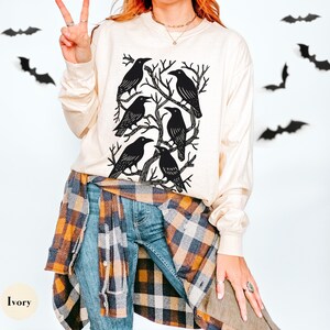May include: Ivory long-sleeve shirt featuring a black graphic of six crows perched on tree branches. The shirt is paired with blue jeans and a plaid shirt tied around the waist. Black bat cutouts decorate the background.