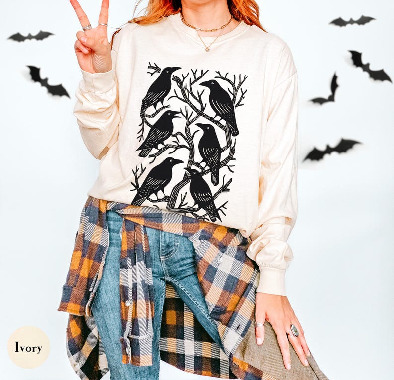 May include: Ivory long-sleeve shirt featuring a black graphic of six crows perched on tree branches. The shirt is paired with blue jeans and a plaid shirt tied around the waist. Black bat cutouts decorate the background.