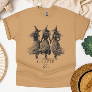 May include: An old gold t-shirt featuring a dark brown graphic of three figures wearing pointed hats and long dresses, holding hands. Below the illustration, the text reads: ESCAPED SINCE 1692. The shirt has short sleeves and a crew neckline.