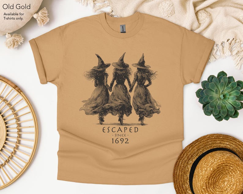 May include: An old gold t-shirt featuring a dark brown graphic of three figures wearing pointed hats and long dresses, holding hands. Below the illustration, the text reads: ESCAPED SINCE 1692. The shirt has short sleeves and a crew neckline.