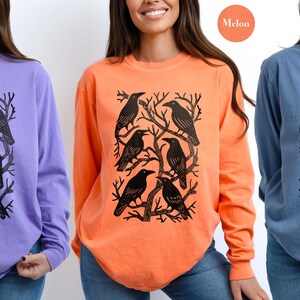 May include: Long-sleeved shirts in violet, melon, and blue jean colors. Each shirt features a black graphic of crows perched on tree branches. The shirts are made of a soft material and are suitable for casual wear.