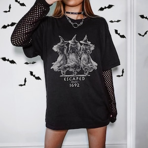 May include: A black t-shirt featuring a white graphic of three witches in pointed hats and long dresses, with the text "ESCAPED SINCE 1692" printed below. The shirt is worn over a black long-sleeved fishnet top, and a silver chain necklace is visible.
