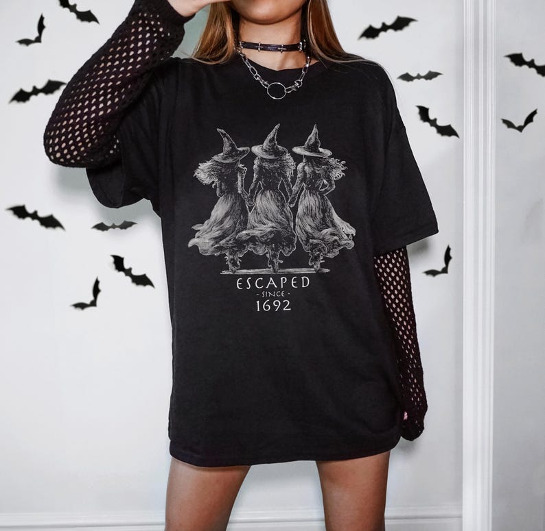 May include: A black t-shirt featuring a white graphic of three witches in pointed hats and long dresses, with the text "ESCAPED SINCE 1692" printed below. The shirt is worn over a black long-sleeved fishnet top, and a silver chain necklace is visible.