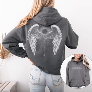 May include: A charcoal gray hoodie featuring a detailed white angel wing design on the back. The design includes a central sunburst and a vertical line with cross-like symbols. The hoodie has a drawstring hood and a kangaroo pocket.