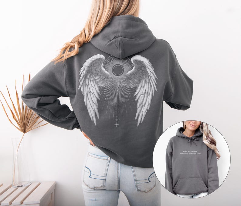 May include: A charcoal gray hoodie featuring a detailed white angel wing design on the back. The design includes a central sunburst and a vertical line with cross-like symbols. The hoodie has a drawstring hood and a kangaroo pocket.