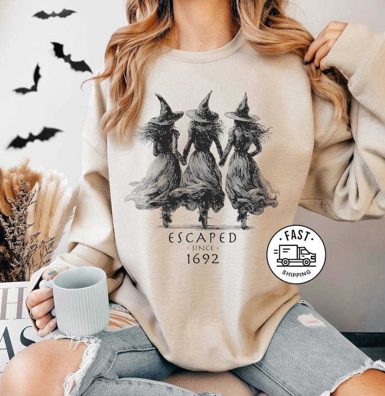 May include: A beige crewneck sweatshirt featuring a black graphic of three witches in pointed hats holding hands, with the text "ESCAPED SINCE 1692" printed below the figures. The design is centered on the chest of the long-sleeved garment.