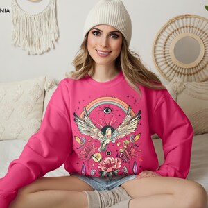 May include: A pink sweatshirt with a colorful graphic print of a bird with wings spread, a rainbow, an eye, and flowers. The graphic is surrounded by stars.