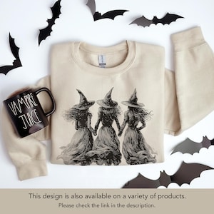 May include: A beige sweatshirt featuring a black and white illustration of three witches holding hands. A black mug with "VAMPIRE JUICE" text sits to the left. Black bat cutouts decorate the background, creating a Halloween theme.