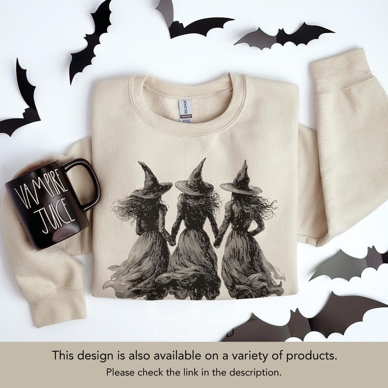 May include: A beige sweatshirt featuring a black and white illustration of three witches holding hands. A black mug with "VAMPIRE JUICE" text sits to the left. Black bat cutouts decorate the background, creating a Halloween theme.