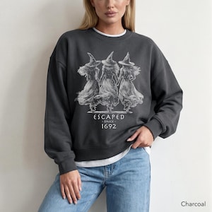 May include: A charcoal gray crewneck sweatshirt featuring a white graphic print of three figures wearing pointed hats and long dresses. Below the figures, the text reads ESCAPED SINCE 1692. The sweatshirt has long sleeves and a ribbed neckline.