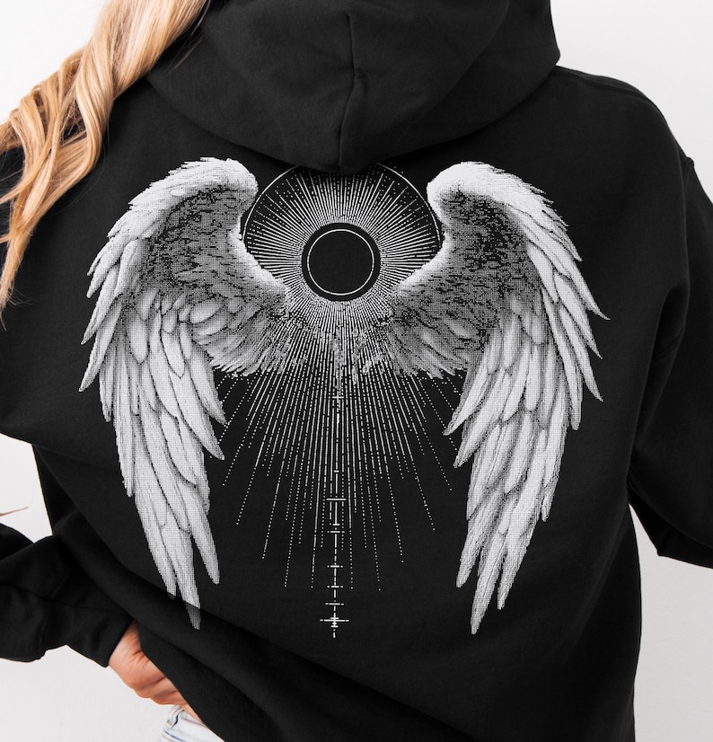 May include: Black hoodie featuring a detailed white angel wing design on the back. The graphic includes a central sunburst element with a circular center and vertical lines. The wings are rendered with intricate feather detailing.