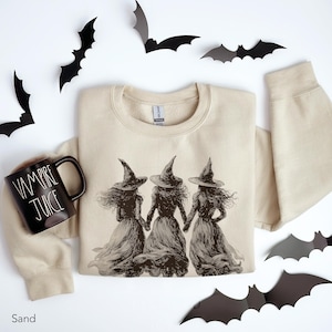 May include: A sand-colored crewneck sweatshirt featuring a dark brown graphic of three witches in pointed hats holding hands. Beside the sweatshirt is a black ceramic mug with the white text Vampire Juice printed on the side.