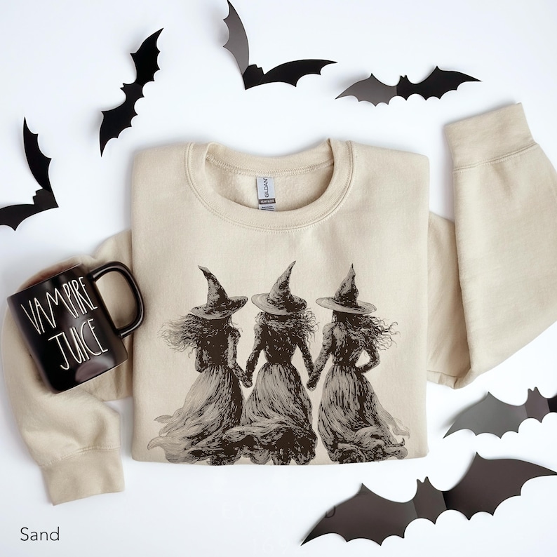 May include: A sand-colored crewneck sweatshirt featuring a dark brown graphic of three witches in pointed hats holding hands. Beside the sweatshirt is a black ceramic mug with the white text Vampire Juice printed on the side.