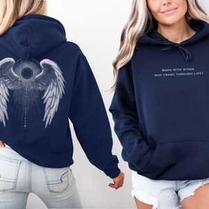 May include: Navy blue hoodie with a large white angel wing design on the back. The front of the hoodie has the text "BORN WITH WINGS WHY CRAWL THROUGH LIFE?" in a light color. The hoodie has a front pocket and a drawstring hood.