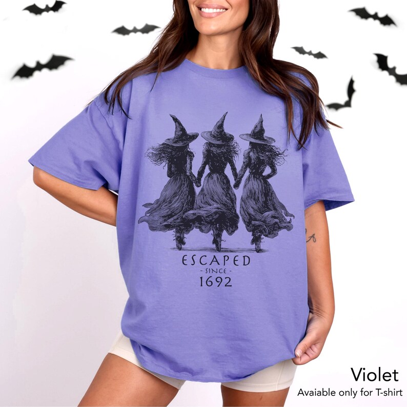 May include: A violet short-sleeved t-shirt featuring a black graphic of three figures in pointed hats and long dresses holding hands. Below the figures, the text reads ESCAPED - SINCE - 1692. The shirt has a crew neckline and a loose, relaxed fit.
