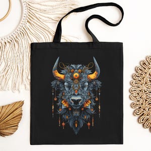 May include: Black tote bag with a colorful illustration of a bull's head with intricate details and a third eye. The bull's head is surrounded by a decorative pattern of feathers and beads.