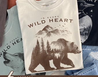 Könnte beinhalten: Eine Auswahl von T-Shirts mit Tiermotiven. Ein cremefarbenes Shirt zeigt eine Bären- und Berg-Grafik mit dem Text "IN EVERY WILD HEART". Weitere Designs sind Wolf, Adler, Hirsch, Pferd und Eistaucher.