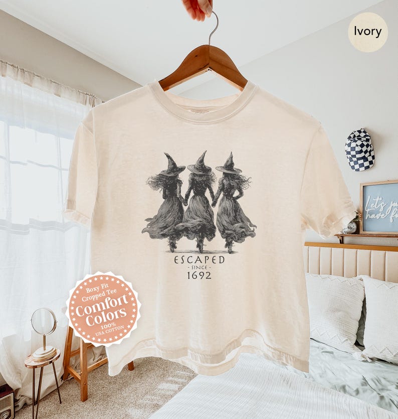 May include: Ivory-colored, boxy fit cropped tee made of 100% USA cotton. The shirt features a graphic of three figures in witch hats and dresses, with the text "ESCAPED - SINCE - 1692". The shirt is hanging on a wooden hanger.