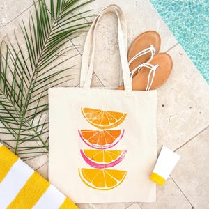 May include: A white canvas tote bag with a design of four stacked orange slices. The slices are in shades of orange, yellow, and pink. The bag has long straps and is lying on a tiled surface next to a towel, sandals, and sunscreen.