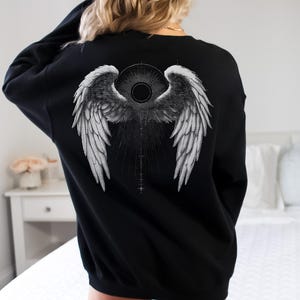 May include: Black sweatshirt featuring a detailed grayscale angel wing design on the back. The graphic includes a central circular element and radiating lines, creating a striking visual effect. The sweatshirt is a classic crew neck style.