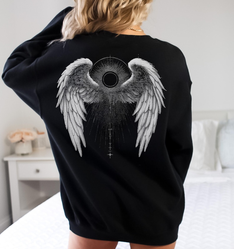 May include: Black sweatshirt featuring a detailed grayscale angel wing design on the back. The graphic includes a central circular element and radiating lines, creating a striking visual effect. The sweatshirt is a classic crew neck style.