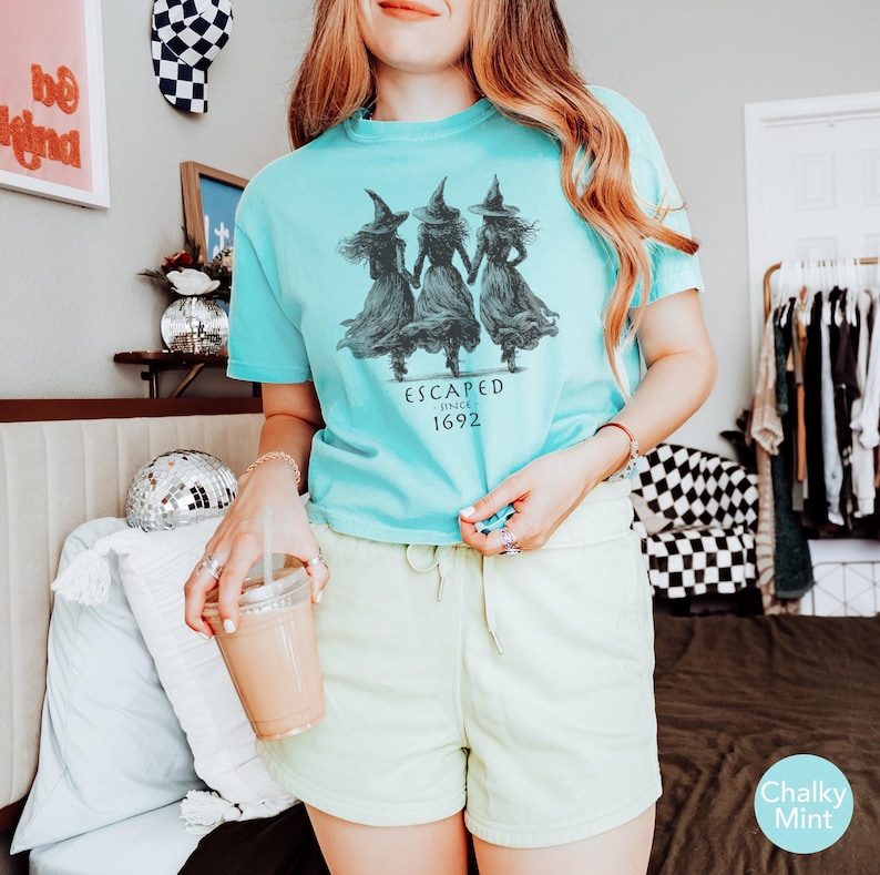 May include: A light blue t-shirt featuring a graphic of three witches holding hands, with the text "ESCAPED SINCE 1692". The shirt is paired with light green shorts. A clear plastic cup with a brown beverage is held in the hand.